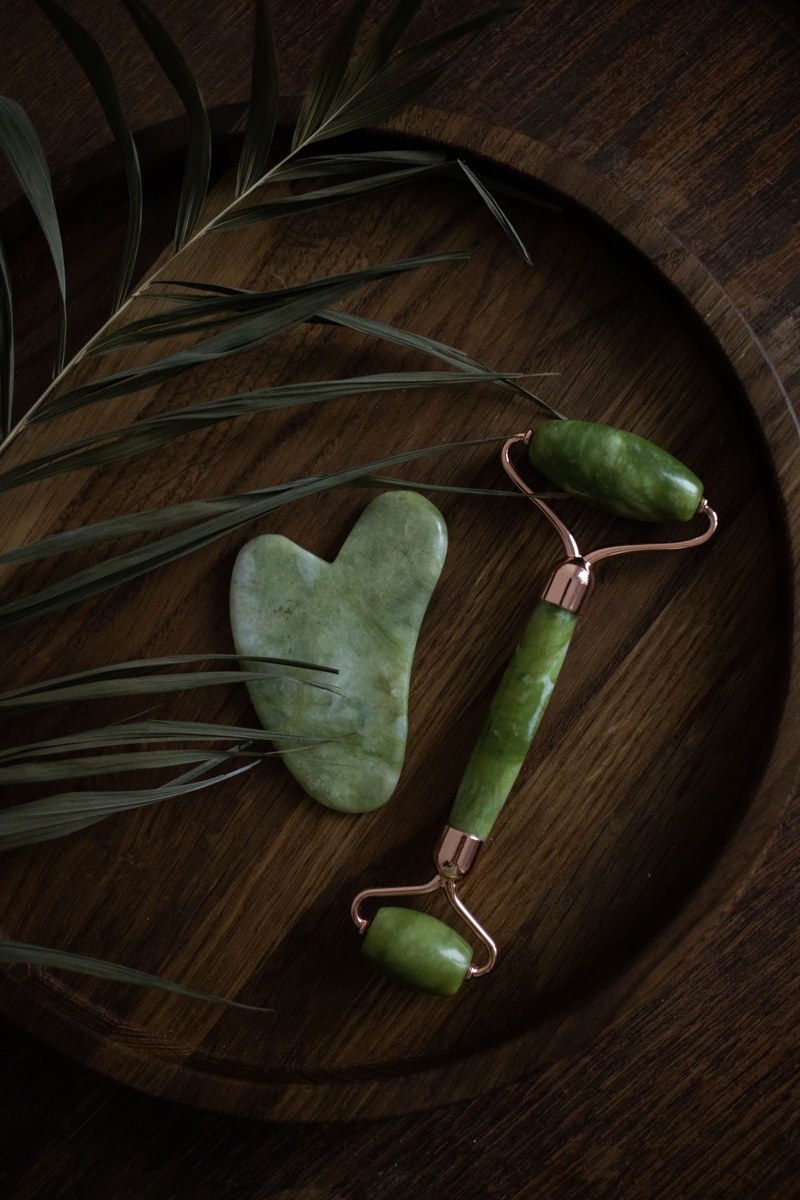 Heart-shaped jade gua sha and dual jade facial roller on a dark wooden tray